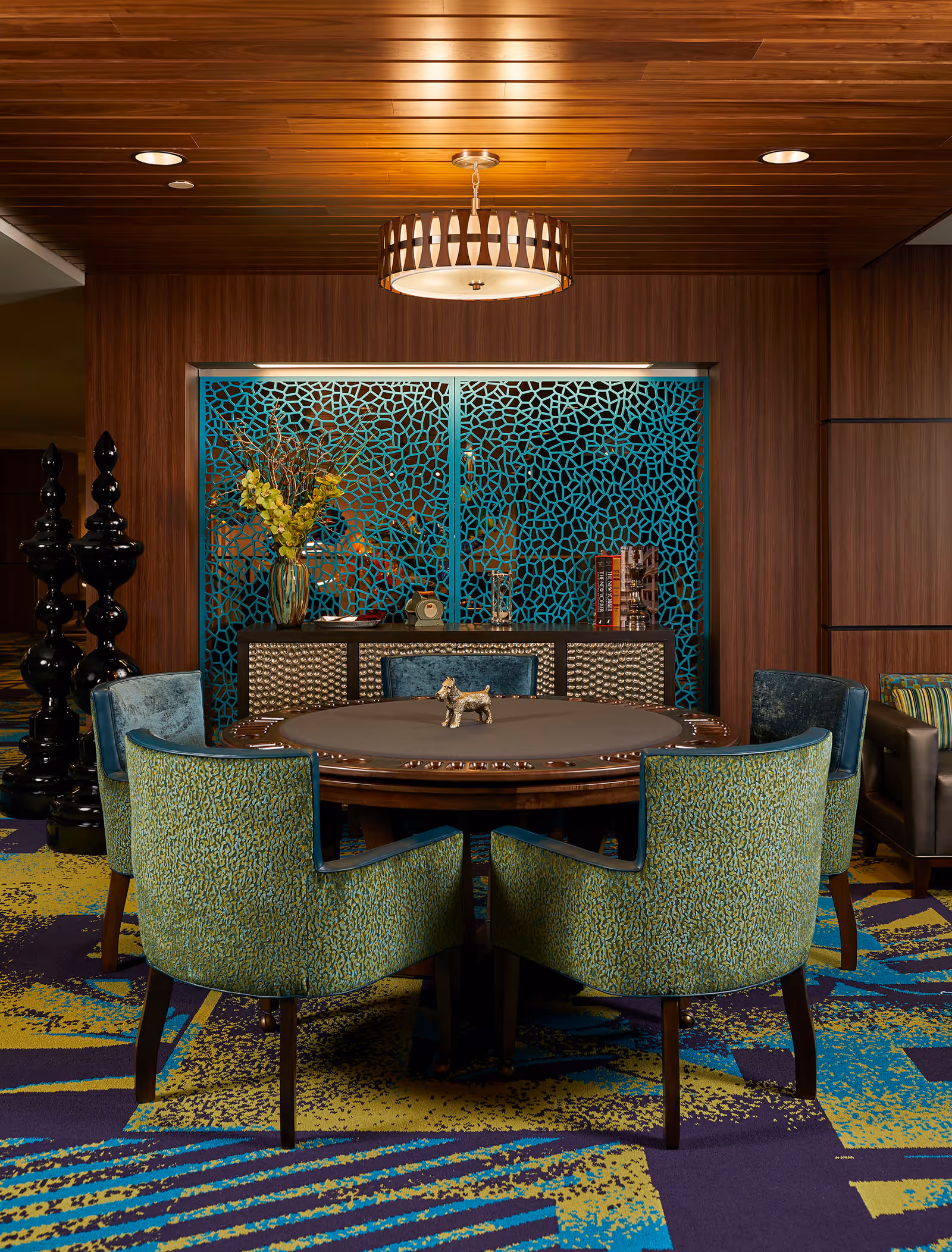 A cozy seating area in a senior living facility featuring a round table surrounded by five green and blue upholstered chairs. The table has a small decorative dog figurine in the center. Behind the table is a wooden cabinet with a decorative blue lattice screen above it, a vase with yellow flowers, and some books. The room has a colorful patterned carpet and a wooden ceiling with a modern circular light fixture.