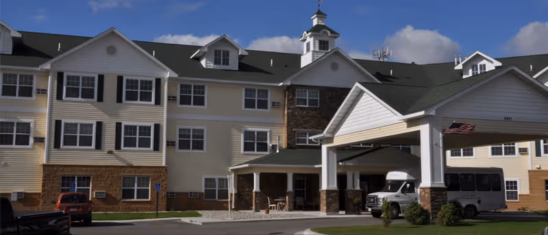 Exterior view of a multi-story senior living facility building with beige siding, white trim, and dark green roof. The entrance features a covered drop-off area with white columns and stone accents. Several vehicles are parked near the entrance under a clear blue sky.