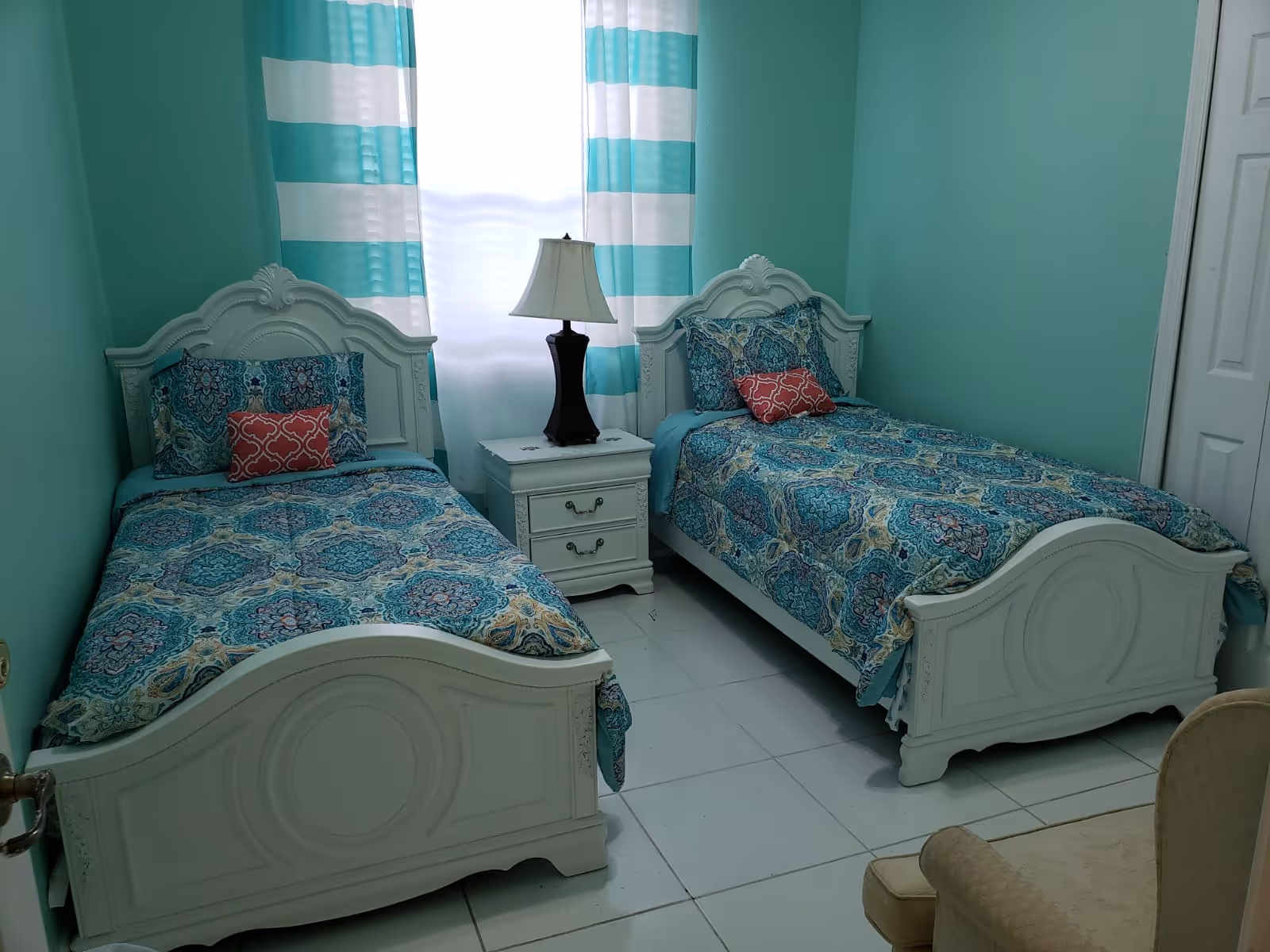 A bedroom with two white twin beds featuring ornate headboards and footboards. Each bed is made with blue patterned bedding and has a small red decorative pillow. Between the beds is a white nightstand with two drawers and a black table lamp. The walls are painted teal, and a window with white and teal striped curtains is centered behind the nightstand. A beige upholstered chair is partially visible in the foreground.