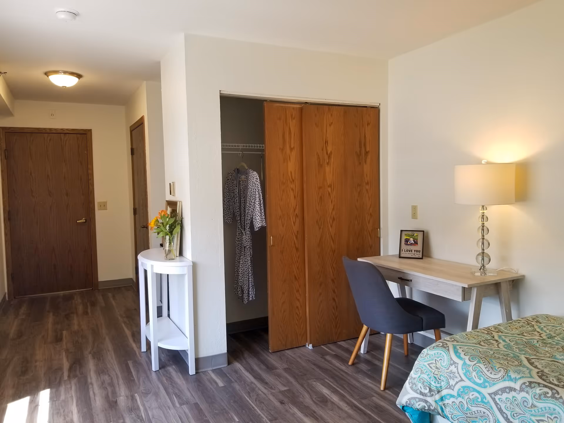 Well-lit assisted living bedroom with patterned bedding, a desk and lamp, an open closet with hanging robes, and wood doors and flooring.