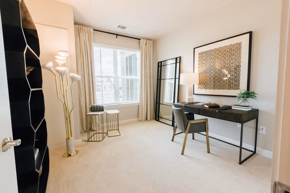 Bright airy home office with a black desk, upholstered chair, floor lamp, large window with curtains, and decorative shelving.