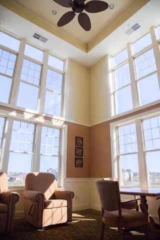 Sunlit double-height common room with tall windows, two upholstered armchairs and a small table with chairs.