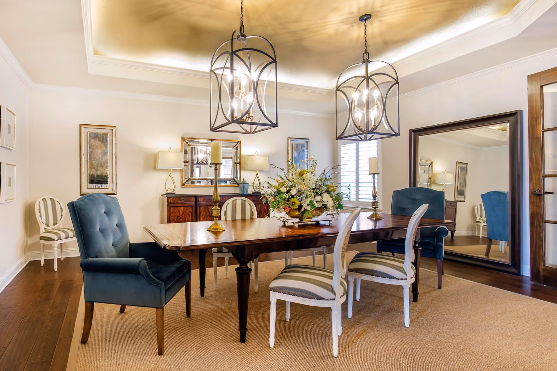 A well-lit dining room with a wooden dining table surrounded by six chairs, including two blue upholstered armchairs and four white chairs with striped cushions. The table is decorated with a floral centerpiece and two candle holders. The room features two large pendant lights hanging from a tray ceiling with gold accents, a large mirror leaning against the wall, framed artwork, and a wooden sideboard with lamps and a mirror above it.