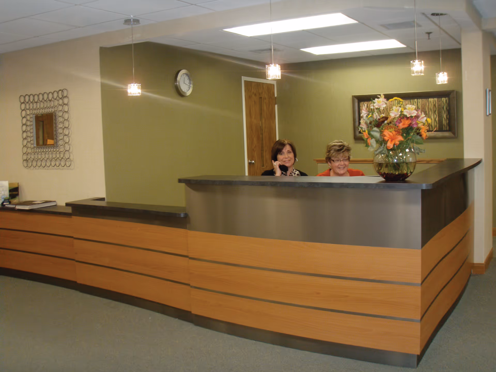 Reception area with a curved wooden and metal desk, two women behind the desk smiling, one on the phone. The wall behind them is green with a clock and a framed picture. A vase with colorful flowers is on the desk.