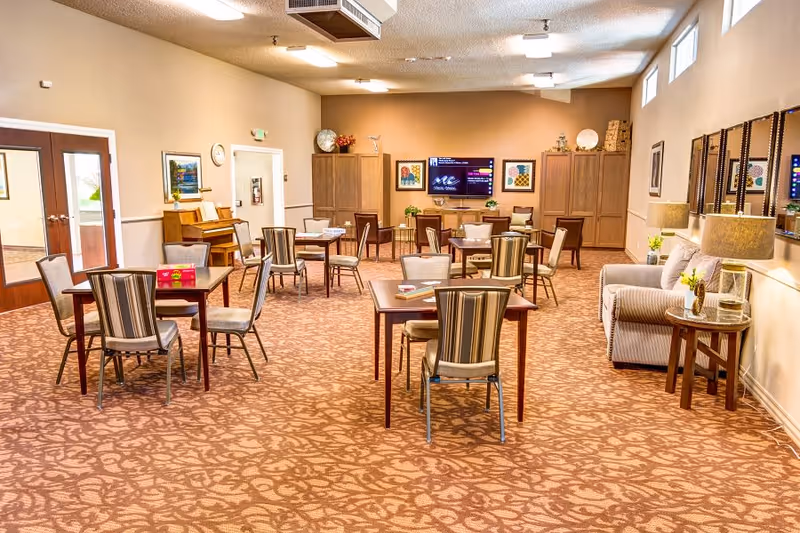 A spacious senior living common area with multiple tables and chairs arranged for socializing or activities. The room features a patterned carpet, a piano against the left wall, a large flat-screen TV mounted on the far wall, and several cabinets. There is a comfortable seating area with a sofa and side tables with lamps on the right side. The room is well-lit with ceiling lights and natural light from high windows.