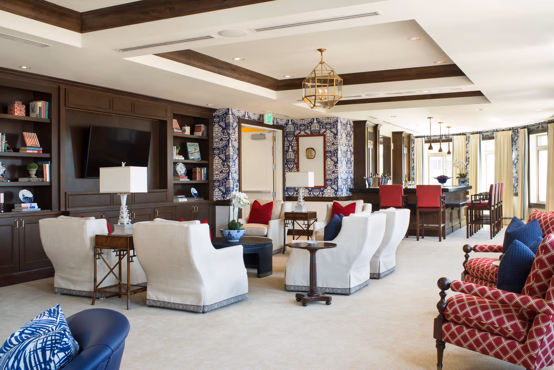 A spacious and elegant living room area featuring white upholstered armchairs arranged around a central coffee table, a dark wood built-in bookshelf with a mounted flat-screen TV, and a bar area with red bar stools in the background. The room has patterned blue and white wallpaper accents, large windows with cream curtains, and decorative pillows in red and blue on the chairs.