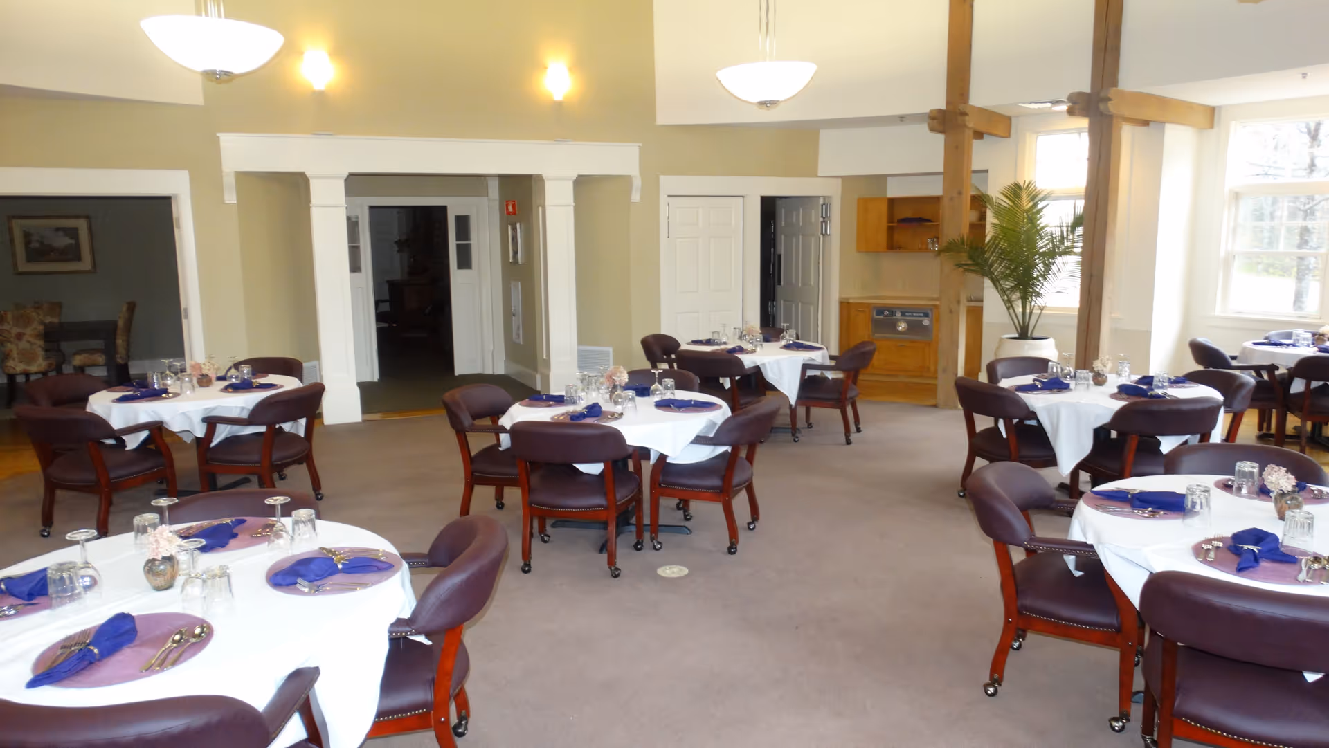 Bright dining room with round tables set with white tablecloths, purple napkins, and wooden chairs.