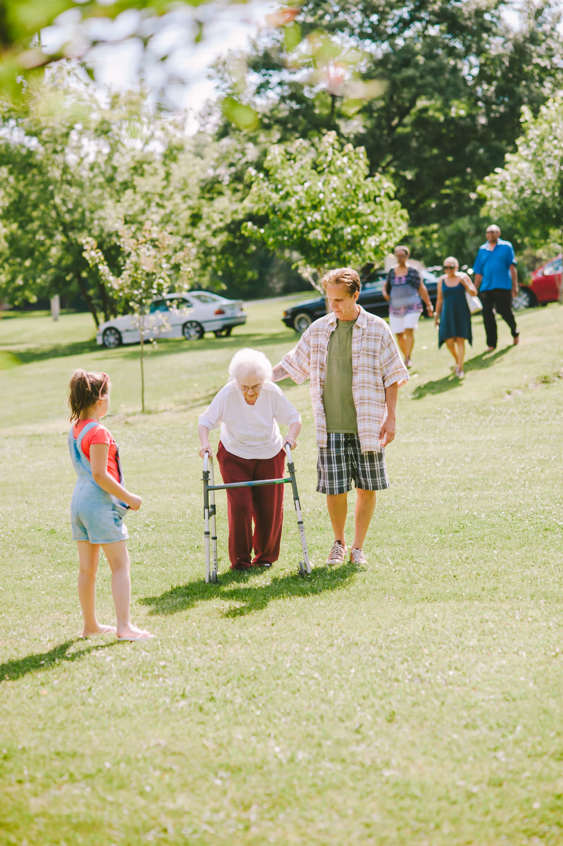 An elderly woman using a walker is being assisted by a man while a young girl stands nearby on a grassy area in a park-like setting. In the background, three other adults walk together among trees and parked cars on a sunny day.