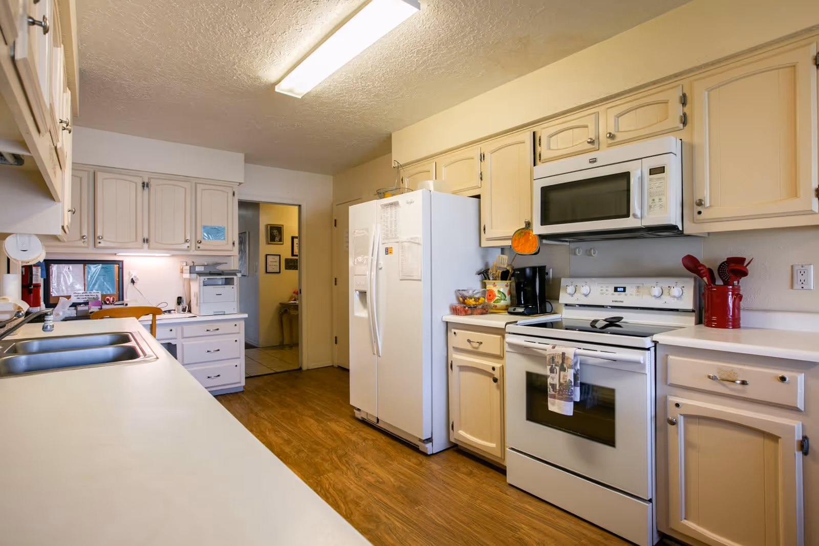 A bright kitchen with cream-colored cabinets and white appliances including a refrigerator, microwave, and stove. The kitchen has a double sink, wooden flooring, and various kitchen utensils and items on the countertops. There is a doorway leading to another room in the background.
