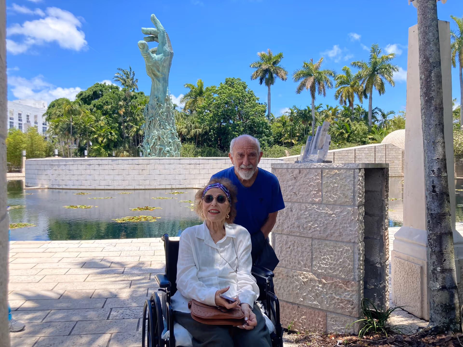 An elderly woman in a wheelchair and an elderly man standing behind her are outdoors near a water feature with lily pads. In the background, there is a large sculpture of an outstretched hand surrounded by greenery and palm trees under a blue sky with some clouds.