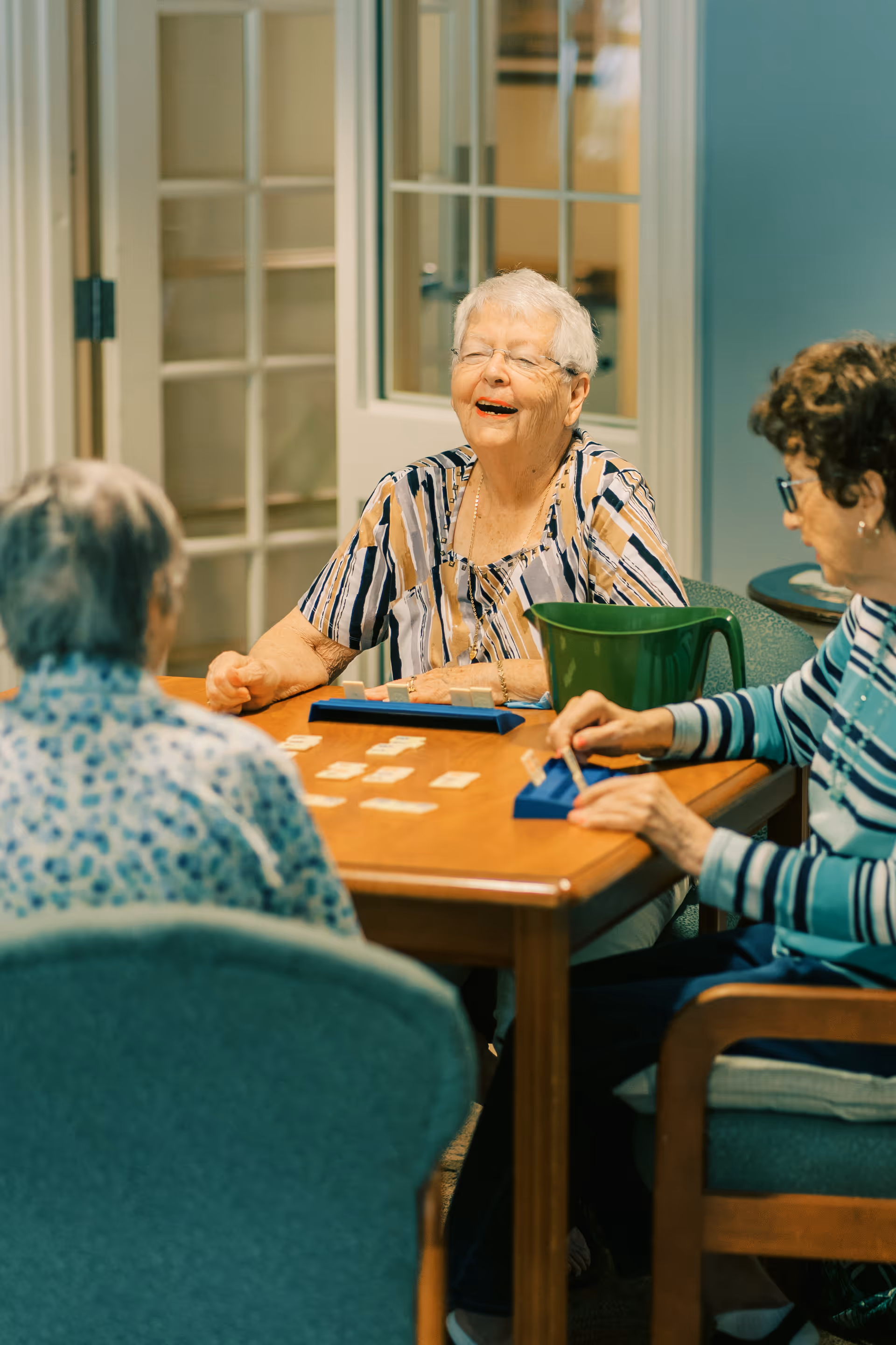 Three elderly women sitting around a wooden table playing a tile-based game, with one woman smiling and laughing. The room has glass-paneled doors in the background and comfortable chairs around the table.
