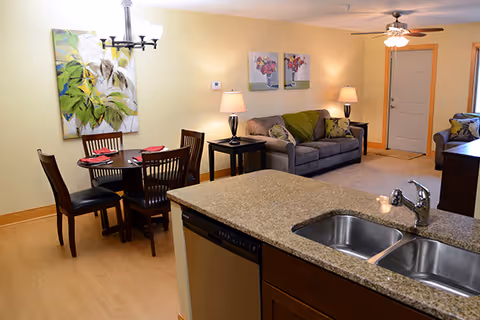 Interior view of a senior living community apartment showing a kitchen island with a double sink in the foreground, a dining area with a round table set for four, and a living room with a gray sofa, side tables with lamps, wall art, and a ceiling fan near the entrance door.