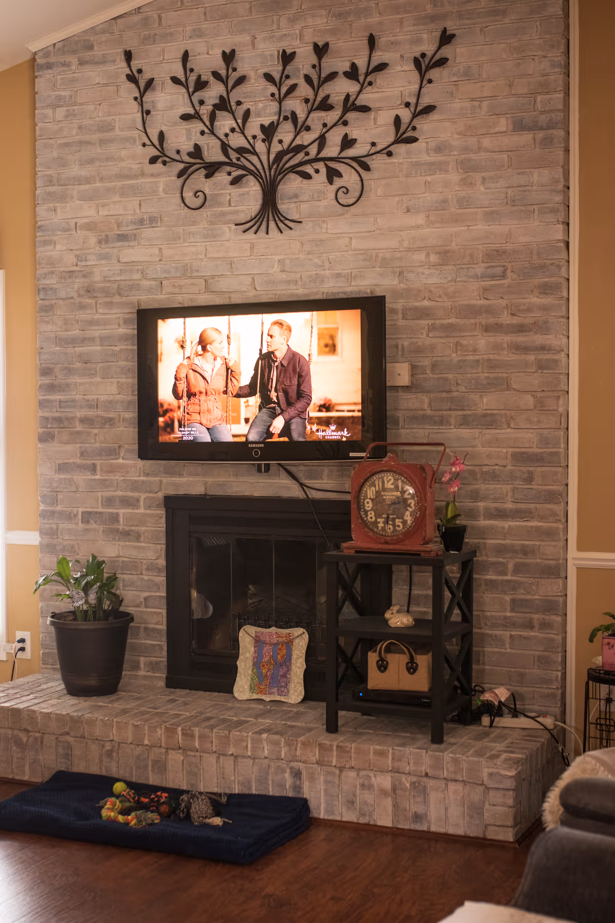 A cozy living room area with a brick fireplace and a mounted flat-screen TV displaying a scene from a Hallmark movie. Above the TV is a decorative black metal wall art piece shaped like branches with leaves. To the right of the fireplace is a small black side table holding a vintage-style clock, a small plant, and a handbag. A potted plant is placed on the left side of the fireplace. On the floor in front of the fireplace is a dark blue mat with some dog toys on it. The room has warm beige walls and wooden flooring.