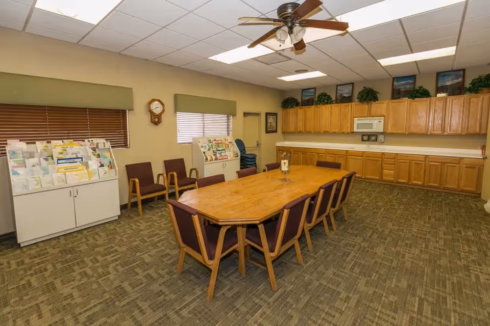 A meeting or dining room with a large wooden table surrounded by chairs with purple cushions. The room has beige walls, carpeted floor, and a ceiling fan with lights. There are wooden cabinets along one wall with a microwave and decorative plants on top. Two windows with green valances and brown blinds are on another wall, along with a clock and racks holding brochures or pamphlets.