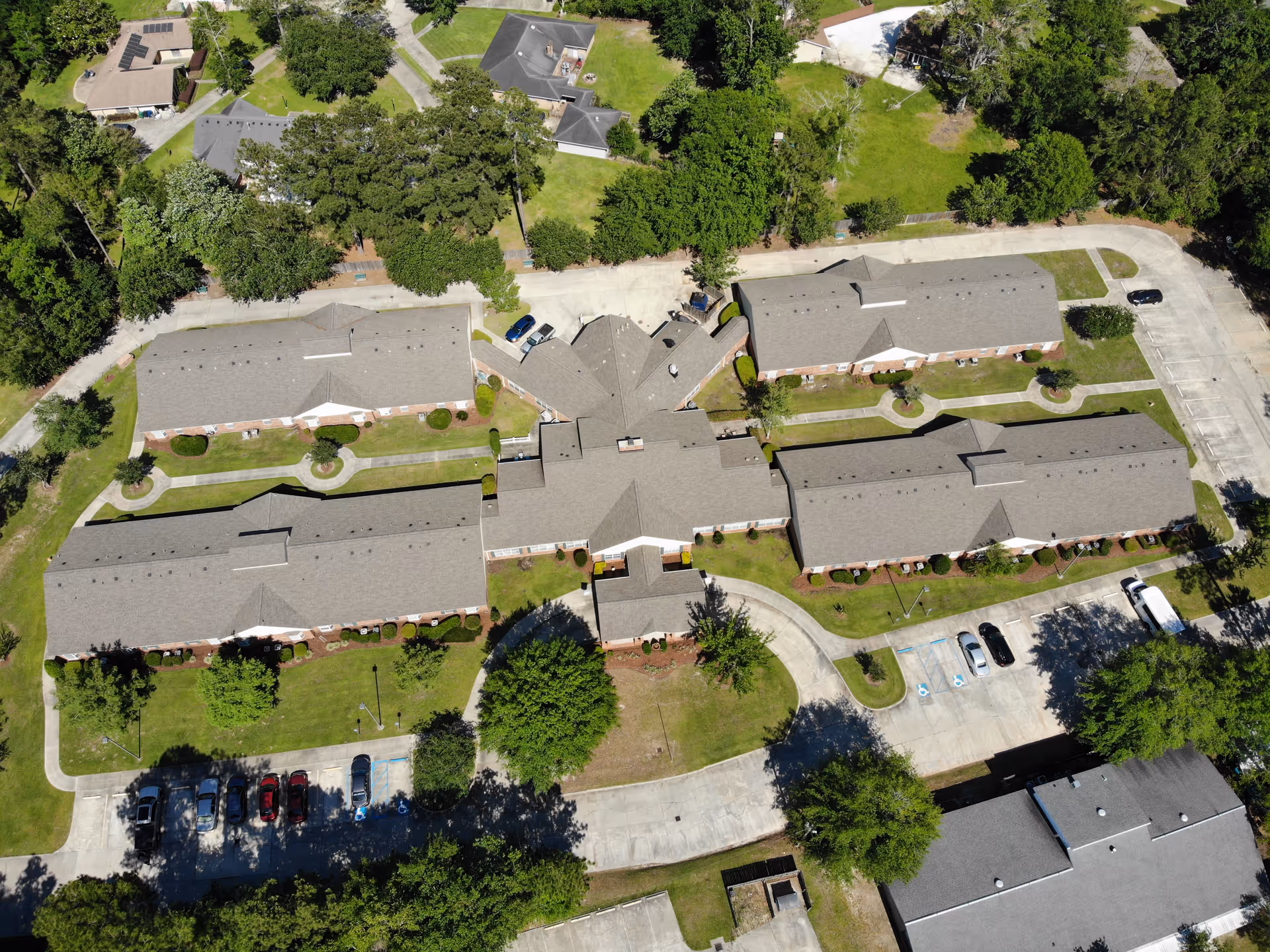 Aerial view of Azalea Estates of Slidell senior living facility showing multiple single-story buildings with gray roofs arranged around a central courtyard with green lawns and trees. Parking lots with cars are visible around the buildings, and surrounding residential houses and trees are seen in the background.