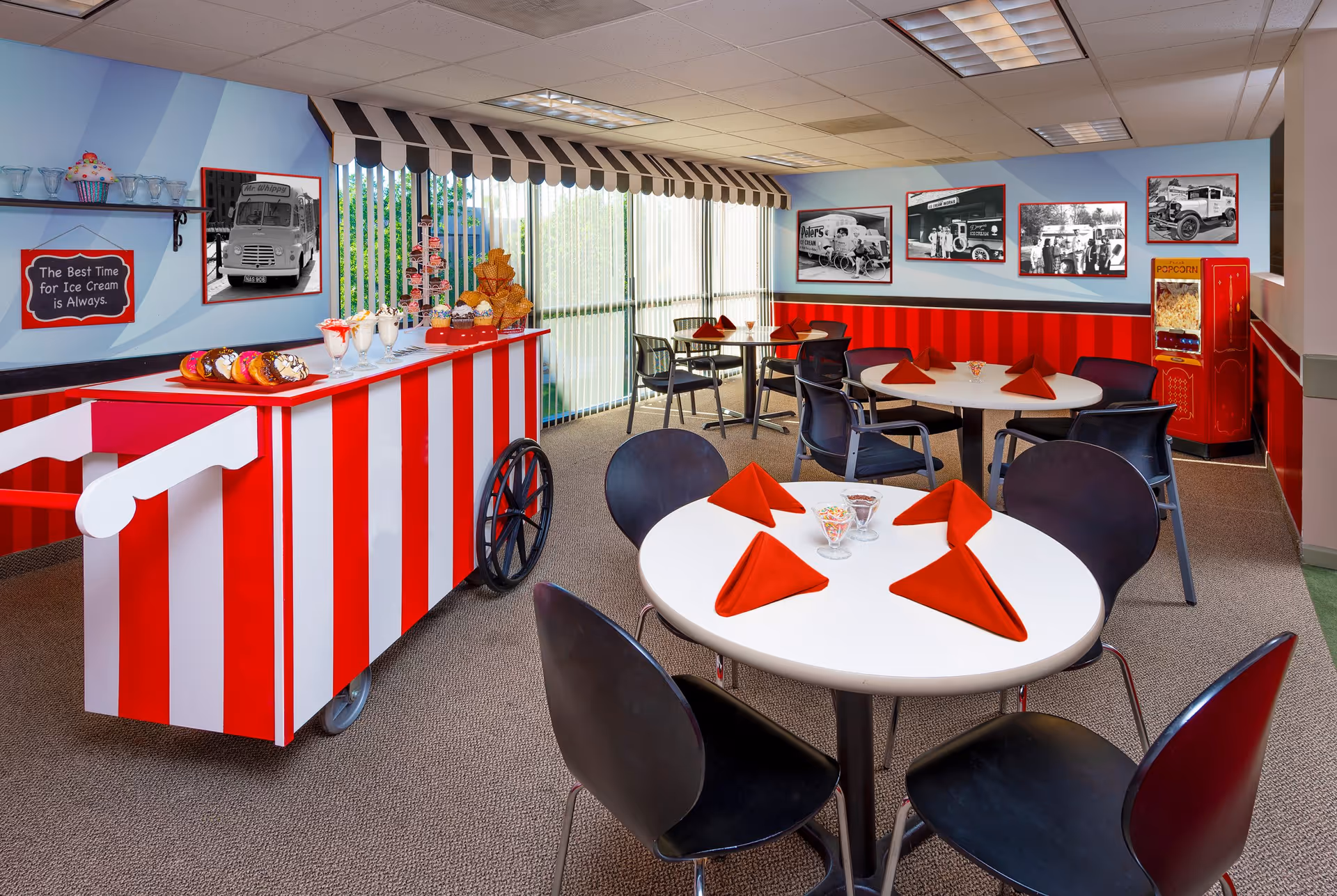 A brightly decorated dining area with a red and white striped ice cream cart displaying donuts, milkshakes, and cupcakes. The room has round tables with black chairs and red folded napkins. The walls are adorned with black and white vintage ice cream truck photos and a sign that reads 'The Best Time for Ice Cream is Always.' There is a popcorn machine in the corner and large vertical blinds covering the windows.
