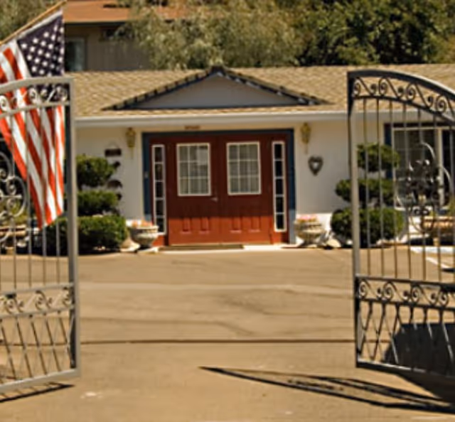 View through open decorative metal gates of the entrance to a single-story building with a brown shingled roof, white walls, and double red doors with glass panels. There are potted plants on either side of the entrance and an American flag on the left side.
