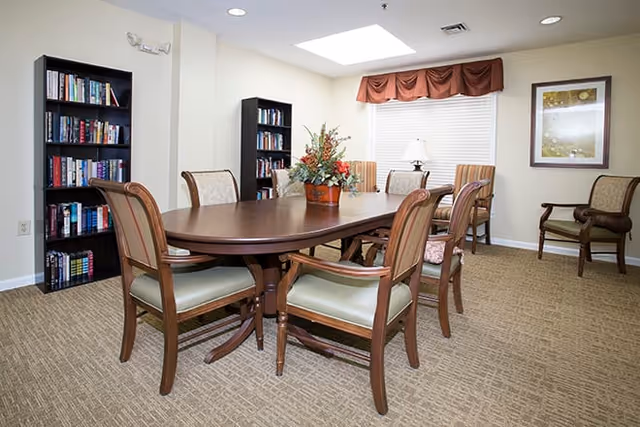 A well-lit room with a large wooden oval table surrounded by six wooden chairs with cushioned seats. There are two tall black bookshelves filled with books against the walls, a window with white blinds and a brown valance, a table lamp on a small side table, and a framed artwork on the wall. The floor is carpeted in a beige pattern.