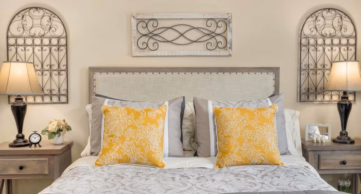 A neatly made bed with a beige upholstered headboard, decorated with two yellow floral-patterned pillows and multiple gray and white pillows. On each side of the bed are wooden nightstands with table lamps, a small clock, a flower vase, and a framed photo. Above the headboard are decorative wrought iron wall hangings.
