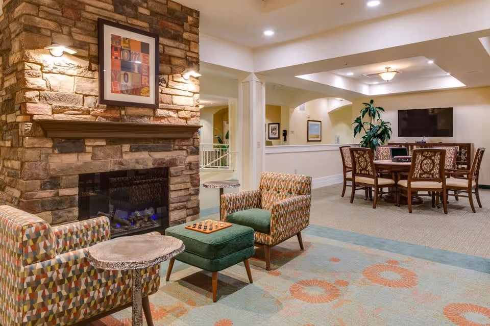 A cozy common area in a senior living facility featuring a stone fireplace with a framed artwork above it, two patterned armchairs with a green ottoman between them, a small round wooden side table, and a carpet with floral patterns. In the background, there is a dining or game table with six chairs and a flat-screen TV mounted on the wall, along with some potted plants and framed pictures on the walls.
