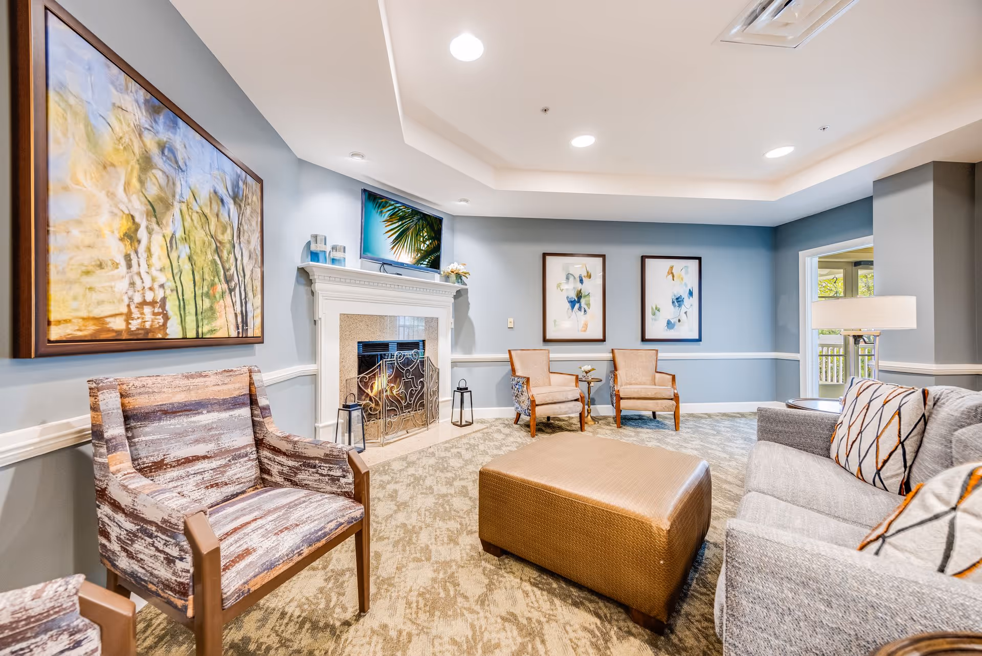 A cozy living room with light blue walls, a fireplace with a decorative screen, a TV mounted above the fireplace, and various seating options including patterned armchairs, a gray sofa with patterned pillows, and a large brown ottoman. The room is decorated with abstract artwork and has a soft carpeted floor.