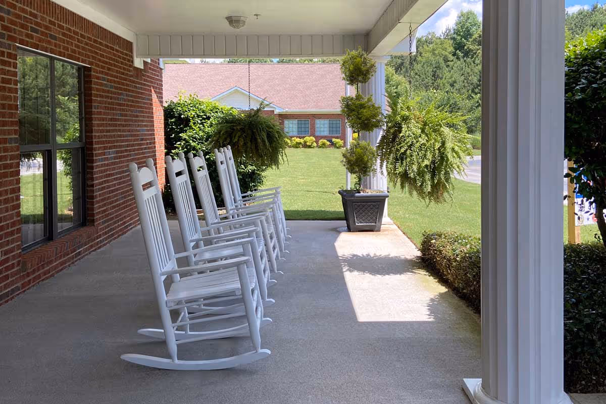 A covered porch area with a row of white wooden rocking chairs lined up along a brick wall. Hanging plants and potted greenery decorate the porch, with a well-maintained lawn and another building visible in the background under a clear blue sky.