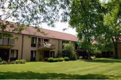 Exterior view of a two-story residential building with balconies, surrounded by green grass, bushes, and large trees providing shade.