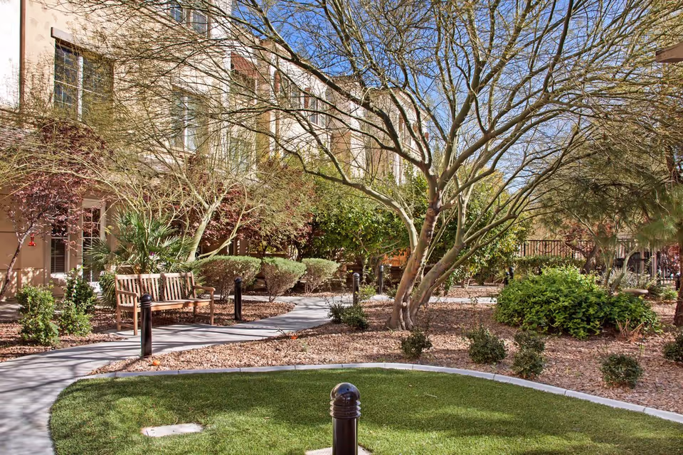 Outdoor garden area at Sunrise of Henderson with a curved concrete pathway, wooden bench, various bushes and trees, and a building with windows in the background under a clear blue sky.