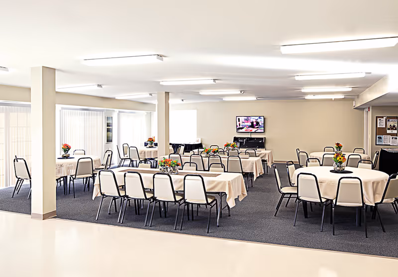 A spacious dining room with multiple tables covered in beige tablecloths, each decorated with colorful flower arrangements. The room has several chairs around the tables, large windows with vertical blinds letting in natural light, and a television mounted on the far wall.