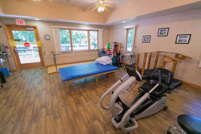 A rehabilitation room with wooden flooring, a blue therapy table with a pillow, parallel bars for walking exercises, a recumbent exercise bike, and various therapy equipment near the windows. The room has large windows letting in natural light and a door with an exit sign above it.