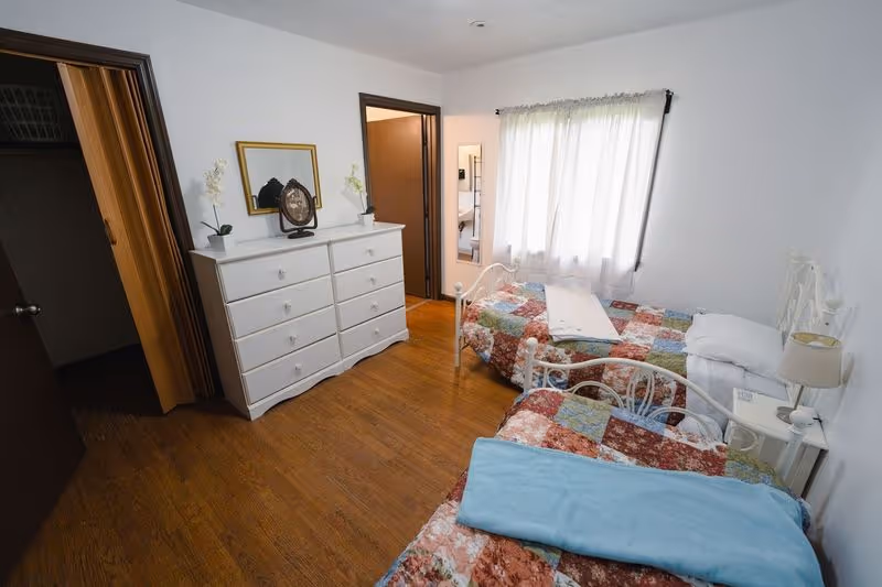 A small bedroom with two single beds covered in colorful patchwork quilts. Each bed has a pillow and a folded blanket on top. There is a white dresser with multiple drawers against one wall, decorated with a mirror, a clock, and small potted plants. A window with white curtains lets in natural light. The floor is wooden, and there are two open doorways visible, one leading to another room and the other to a closet.