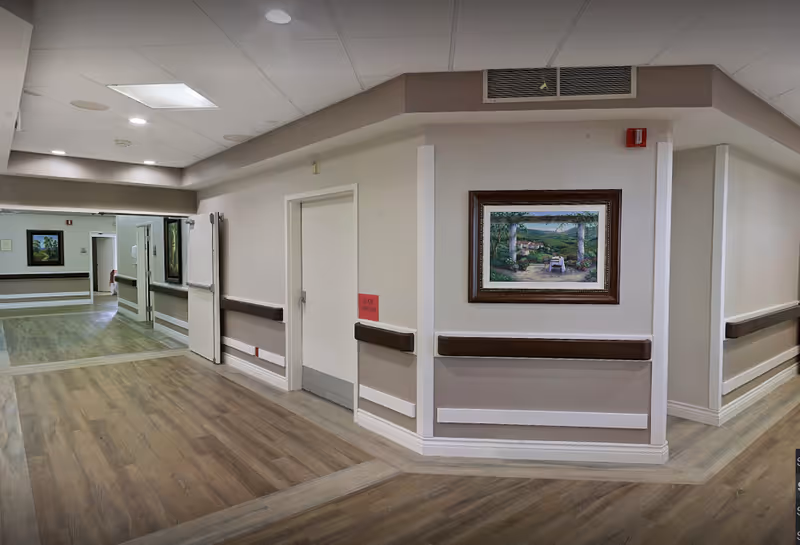 Well-lit hallway in an assisted living facility with wood-look floors, wall handrails, and framed artwork.