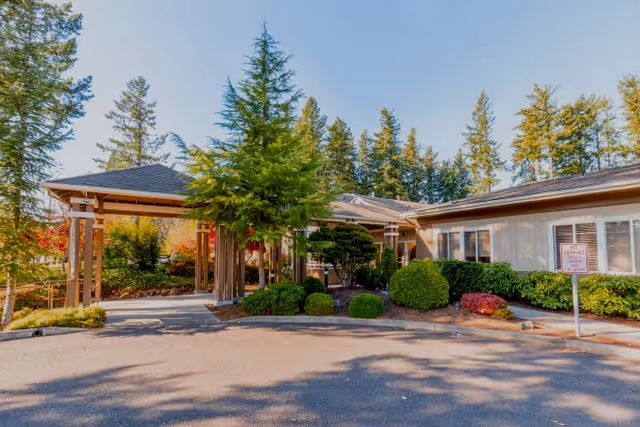 Exterior view of Maple Grove Memory Care facility showing a single-story building with a covered entrance, surrounded by trees and landscaped bushes under a clear blue sky.