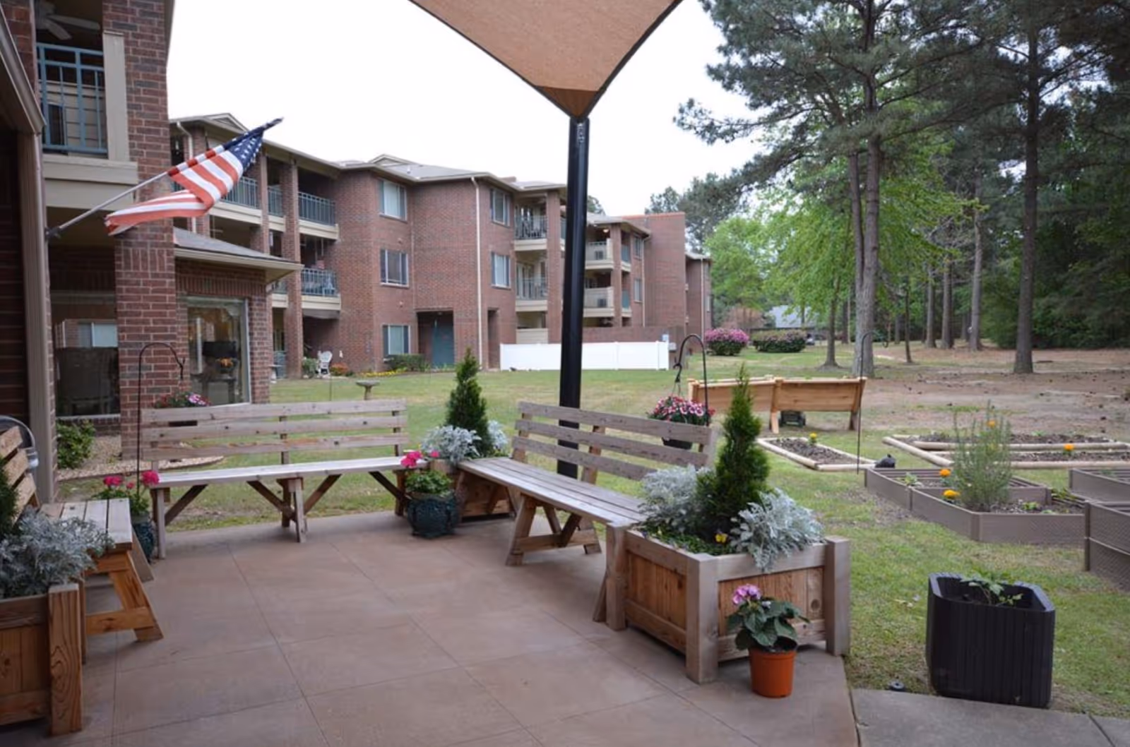 Outdoor patio with wooden benches, planters and raised garden beds in front of a brick apartment building.