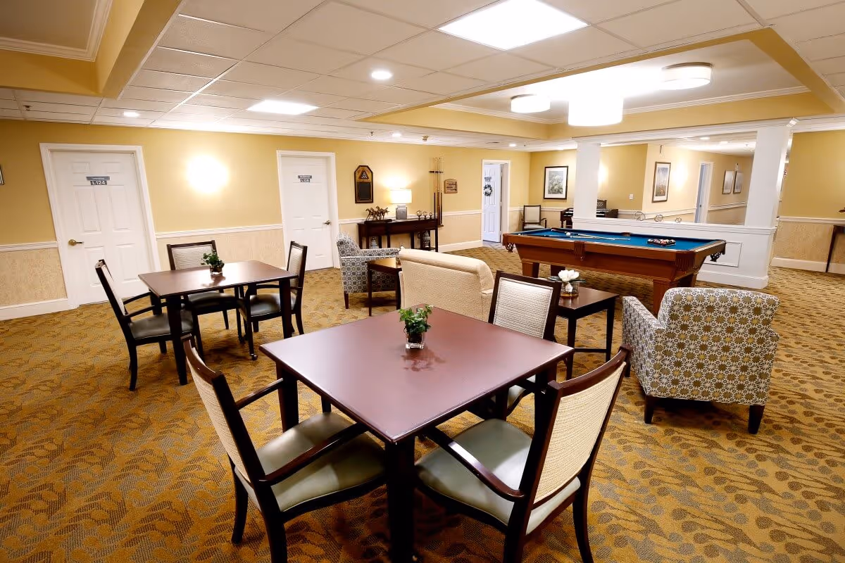 A well-lit common area in a senior living facility featuring multiple square tables with chairs, a seating area with armchairs and a sofa, and a pool table. The walls are painted yellow with white trim, and there are several doors and framed pictures on the walls. The carpet has a patterned design, and the ceiling has recessed lighting and ceiling panels.