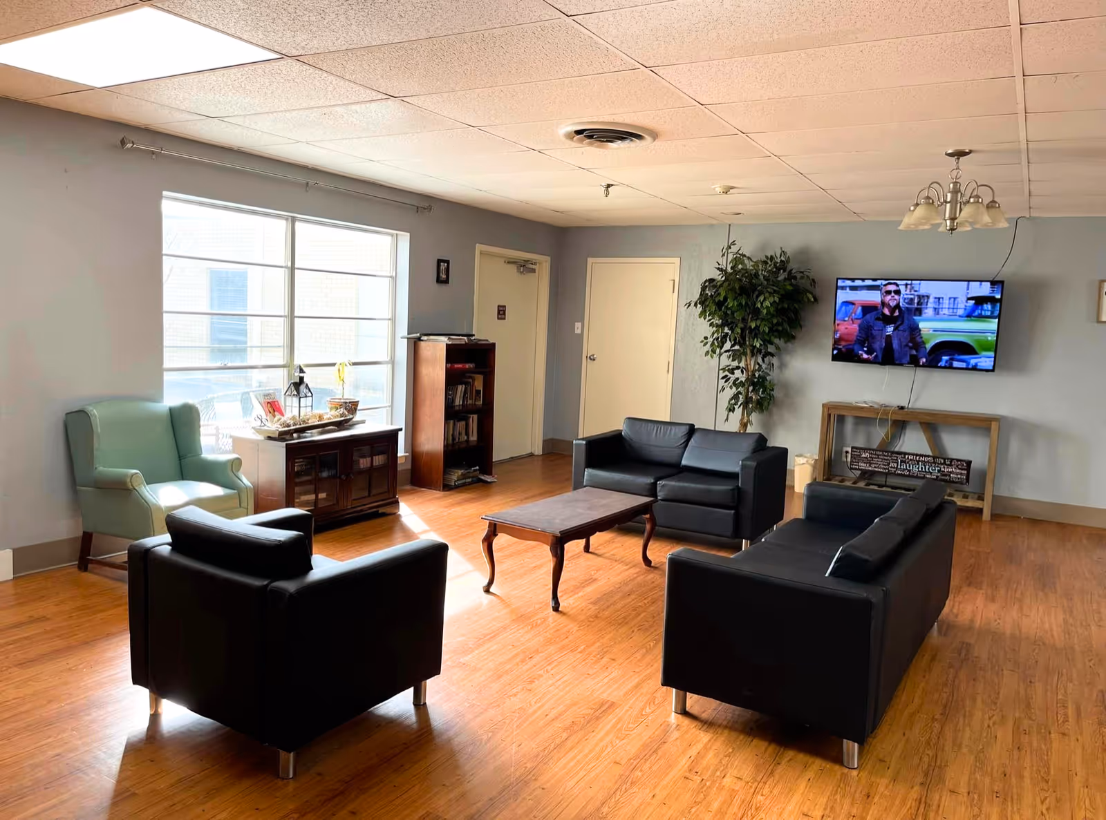 A well-lit communal living room with black leather sofas and chairs arranged around a wooden coffee table, a wall-mounted TV, bookshelf, and large window.