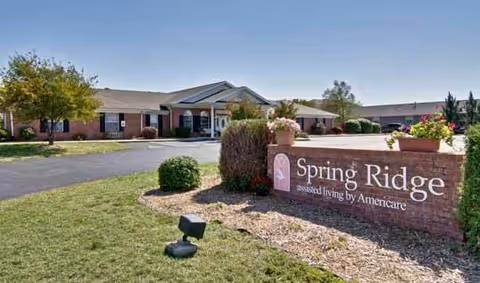 Exterior view of Spring Ridge Assisted Living facility showing a single-story brick building with a covered entrance, surrounded by landscaping including bushes, flowers, and a lawn. A brick sign in the foreground reads 'Spring Ridge assisted living by Americare.'