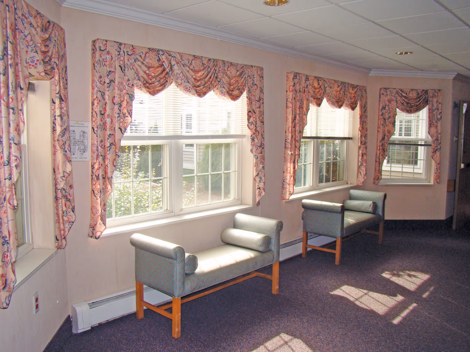 A hallway with three windows covered by floral-patterned curtains. Below each window is a cushioned bench with wooden legs and cylindrical pillows. Sunlight streams through the windows, casting shadows on the carpeted floor.