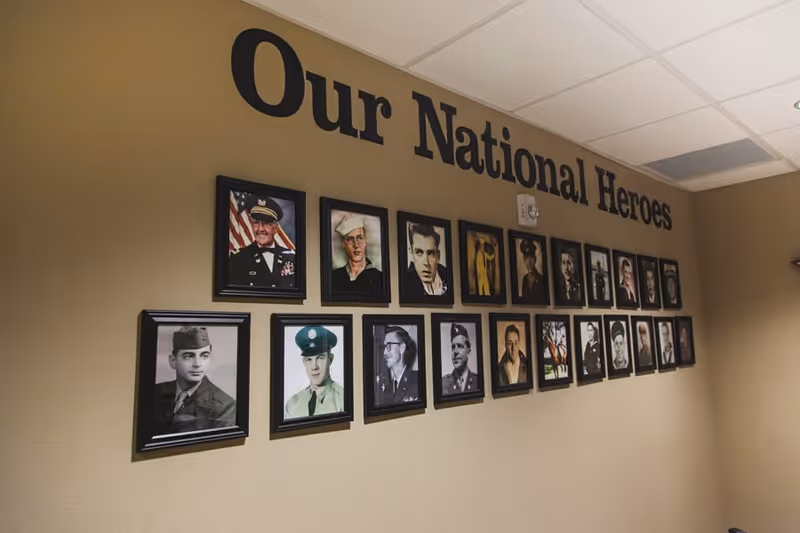 A wall display titled 'Our National Heroes' featuring framed photographs of military veterans in uniform, arranged in two rows on a beige wall under a white ceiling with fluorescent lighting.
