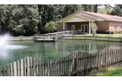 A peaceful outdoor scene at Suwannee Valley Nursing Center featuring a pond with a water fountain, surrounded by a wooden fence. In the background, there is a single-story brick building with a covered porch and a wheelchair ramp, set against a backdrop of trees.