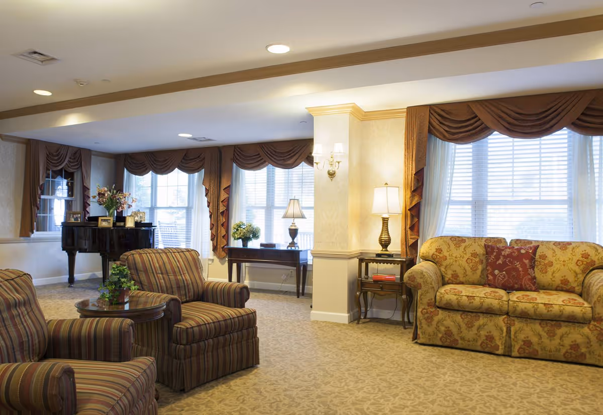 A cozy living room area with two striped armchairs and a round wooden table between them, a floral patterned sofa with a red decorative pillow, a side table with a lamp, a console table with a lamp and flowers, large windows with brown drapes, and a black grand piano in the background.