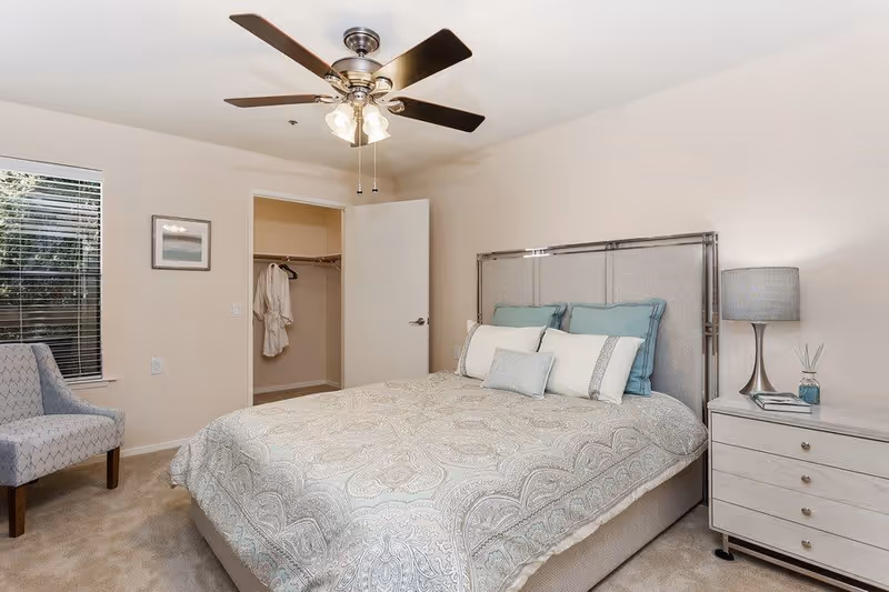 A bedroom with a neatly made bed featuring a patterned bedspread and multiple pillows. There is a ceiling fan with lights above the bed. To the right of the bed is a nightstand with a lamp, a book, and a small decorative item. An open door reveals a walk-in closet with a hanging robe. A window with blinds and a patterned chair are on the left side of the room.