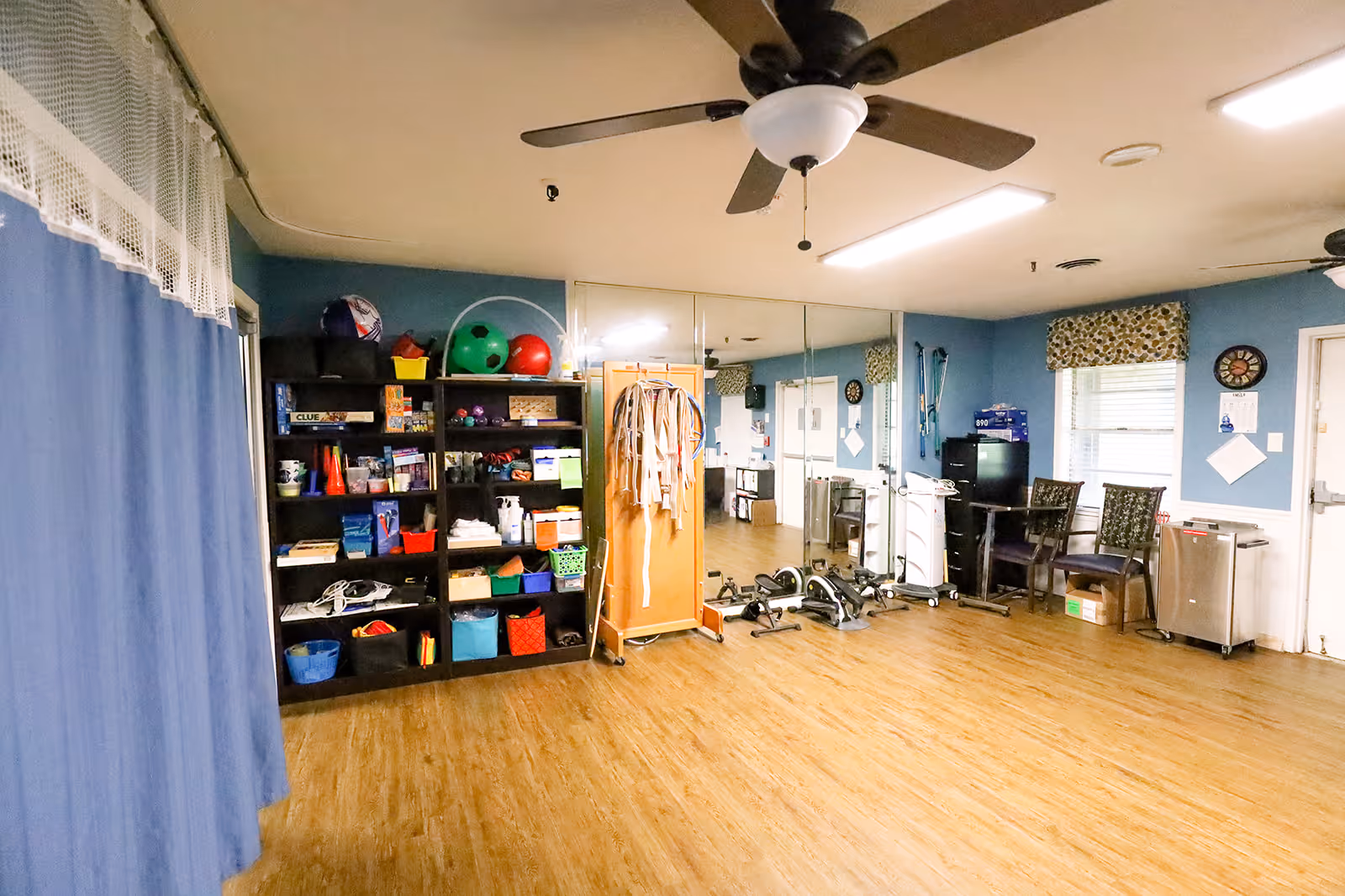 Activity room with shelves of games and supplies, exercise equipment, a mirrored wall, chairs and ceiling fans.