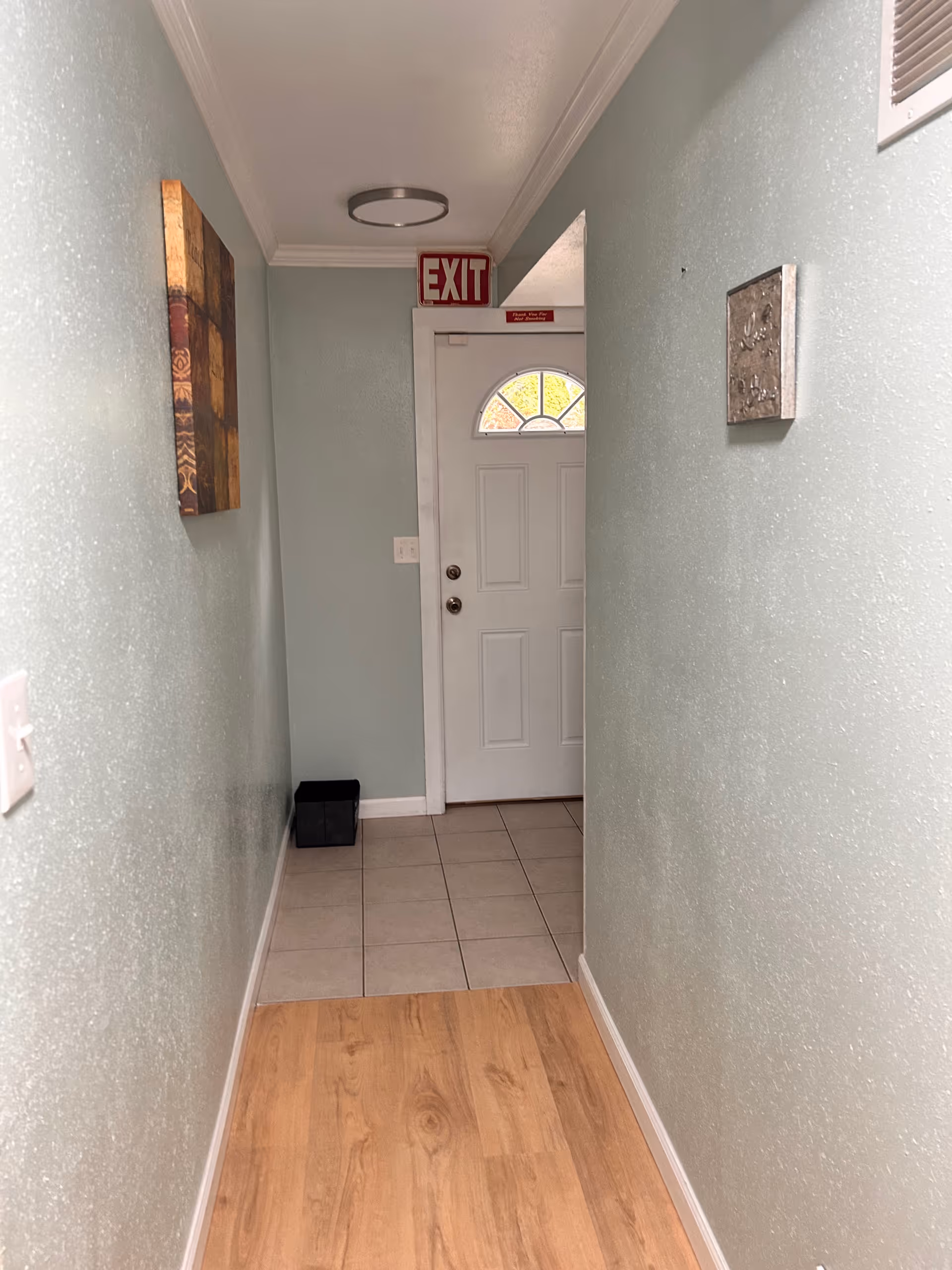Narrow interior hallway with light green walls and wood flooring leading to a white exit door topped by an red EXIT sign.