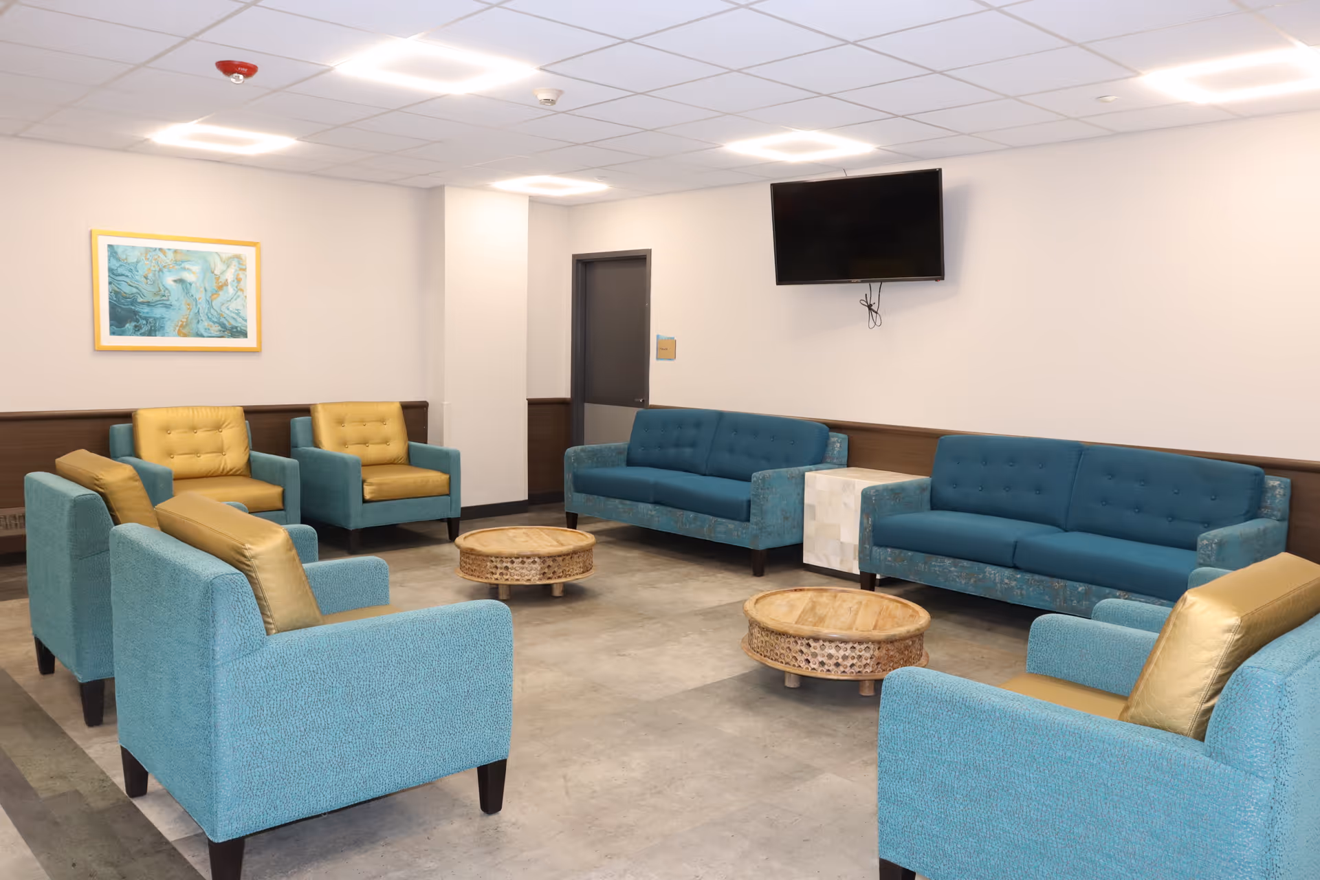 A seating area in a nursing and rehabilitation center with two blue sofas, four blue armchairs with yellow cushions, two round wooden coffee tables, a wall-mounted flat screen TV, and a framed abstract painting on the wall.