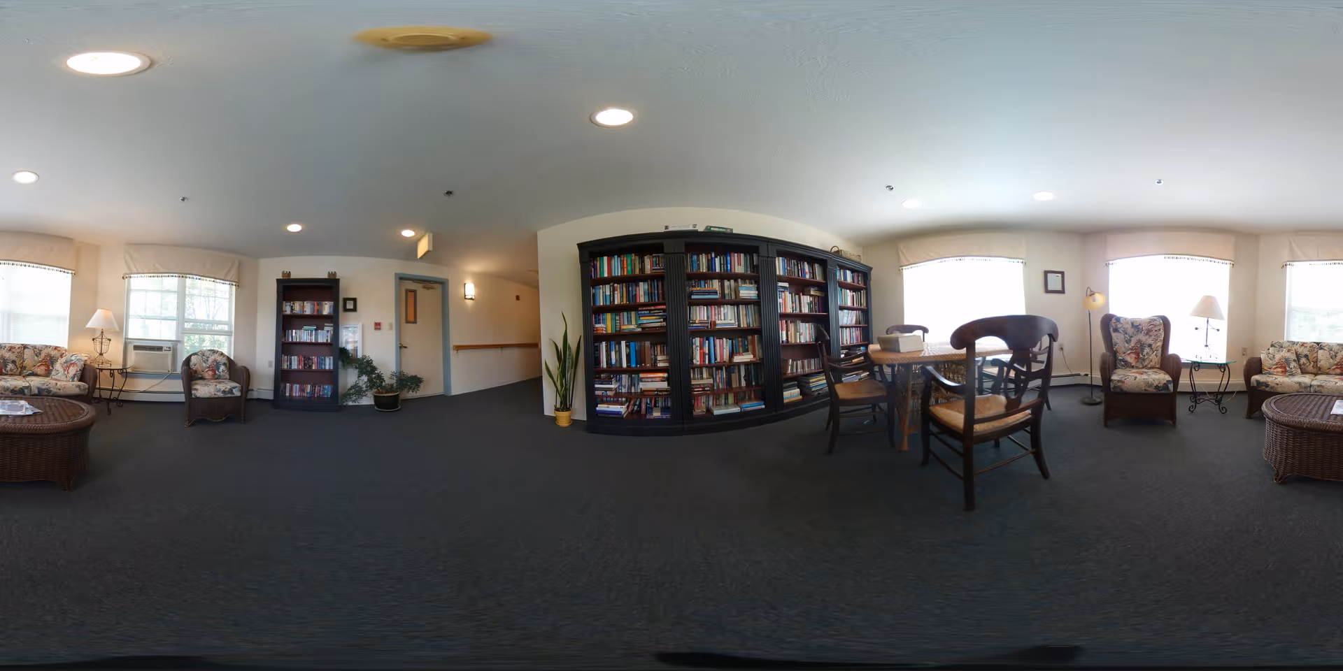 A spacious senior living community common area with carpeted floor, multiple windows letting in natural light, floral upholstered armchairs and sofas, a wicker coffee table, a tall bookshelf filled with books, a small round table with wooden chairs, and potted plants. The walls are light-colored and the ceiling has recessed lighting.