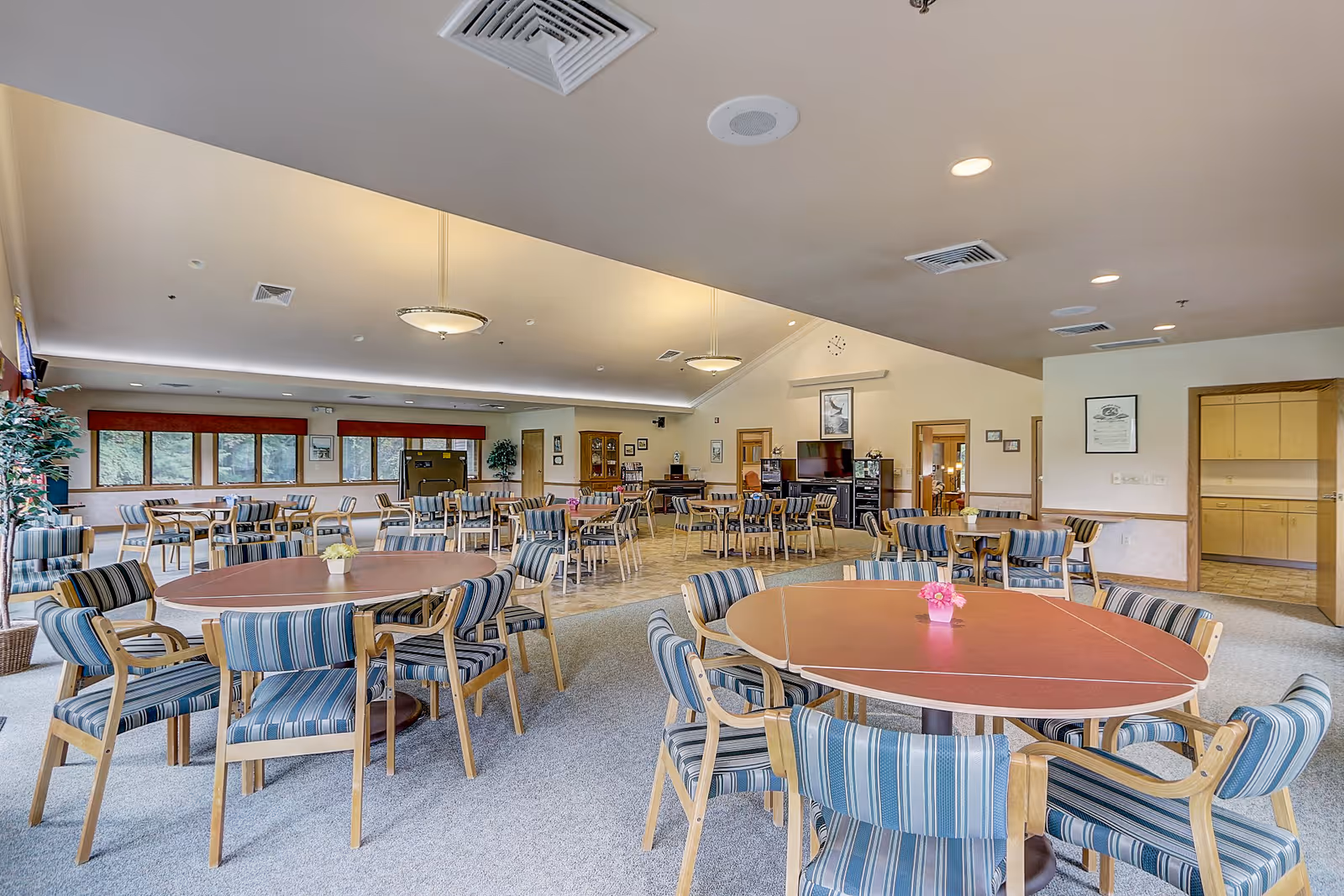 A spacious dining room with multiple round tables surrounded by striped cushioned chairs. The room has large windows with red valances, light-colored walls, and ceiling lights. There are plants and framed pictures on the walls, and a television is visible in the background.