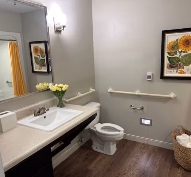 A clean and accessible bathroom with a white sink and countertop, a toilet with grab bars on the walls, a basket with rolled towels, a framed sunflower artwork on the wall, and a vase of yellow tulips on the counter. The floor is wood-style, and there is a yellow shower curtain visible in the mirror reflection.