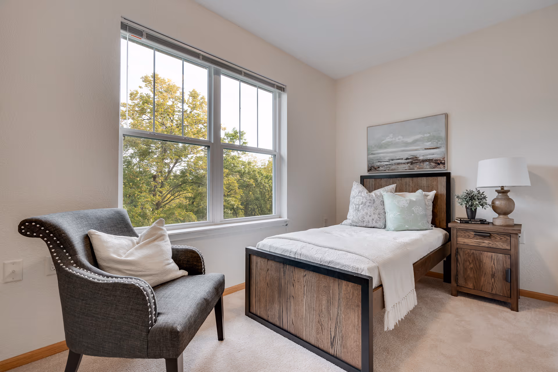 A cozy bedroom with a single wooden bed adorned with white and patterned pillows and a white blanket. Next to the bed is a wooden nightstand with a lamp, a small plant, and books. A gray upholstered chair with a white pillow is positioned near a large window showing green trees outside. A painting hangs above the bed on the light-colored wall.