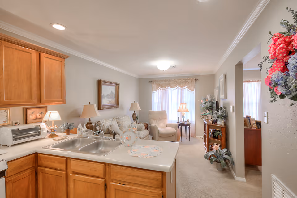 View of a cozy living area from the kitchen in a senior living facility. The kitchen features wooden cabinets, a double sink, and a countertop with a decorative plate and doily. The living room has a sofa, an armchair, two table lamps, a framed painting on the wall, and a window with sheer curtains allowing natural light. There are decorative plants and flowers near the entrance to another room.